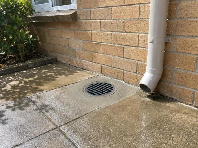 Concrete paving beside a brick wall with a clean round drain grate and downpipe in Pagewood