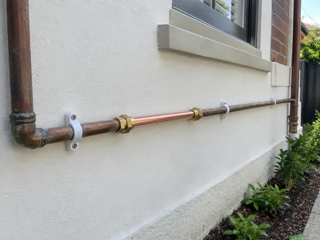 Repaired copper water pipe fixed to a rendered exterior wall beside a window in Kensington